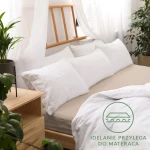 Beżowe prześcieradło jersey z gumką o wymiarach 70x140, leżące na łóżku z kilkoma białymi poduszkami. Zbliżenie na tkaninę, która przylega do materaca. W tle drewniane akcenty i rośliny doniczkowe. Proste tło, które podkreśla produkt.